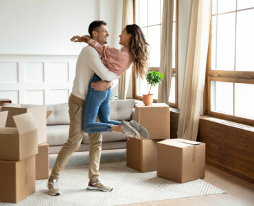 Happy young husband lifting excited wife celebrating moving day with cardboard boxes, proud overjoyed family couple first time home buyers renters owners having fun enjoy relocation, mortgage concept