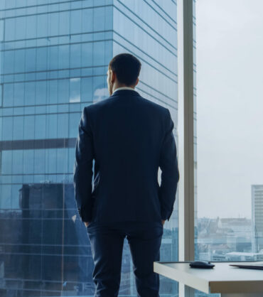 Back View of the Thoughtful Businessman wearing a Suit Standing in His Office, Hands in Pockets and Contemplating Next Big Business Deal, Looking out of the Window. Big City Business District Panoramic Window View.