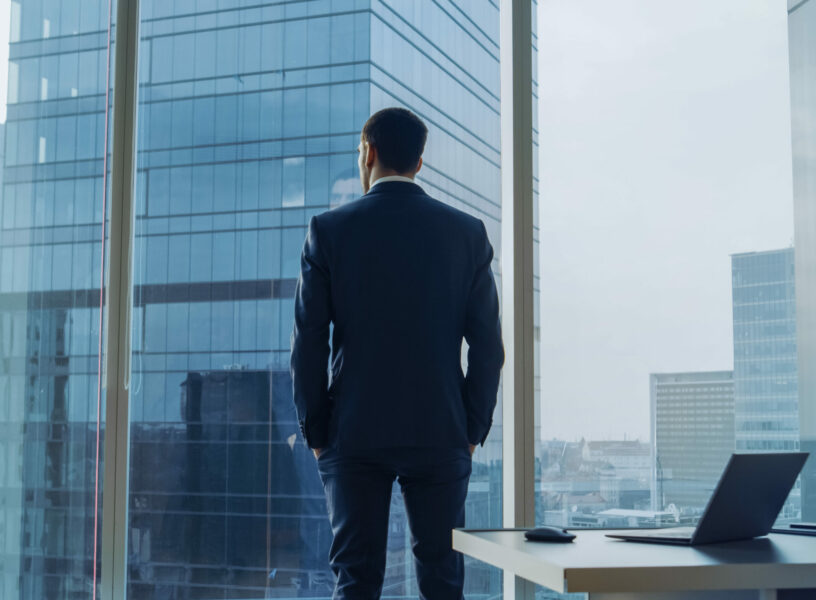 Back View of the Thoughtful Businessman wearing a Suit Standing in His Office, Hands in Pockets and Contemplating Next Big Business Deal, Looking out of the Window. Big City Business District Panoramic Window View. Back View of the Thoughtful Businessman wearing a Suit Standing in His Office, Hands in Pockets and Contemplating Next Big Business Deal, Looking out of the Window. Big City Business District Panoramic Window View.