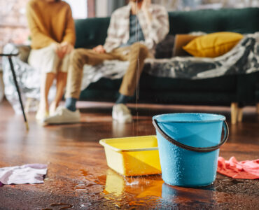 Roof is Leaking, Pipe Rupture at Home: Water Drips into Buckets in Living Room. Angry Couple in Background Calling Insurance Company, Screaming into Phone in Frustration, Trying to Find Plumber