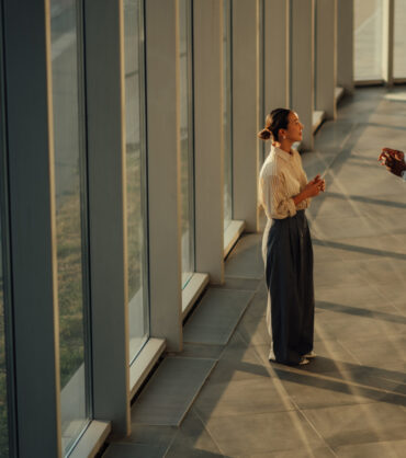 Two businesspeople are having a discussion near a large window in a modern office building, illuminated by warm sunlight