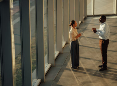 Two businesspeople are having a discussion near a large window in a modern office building, illuminated by warm sunlight