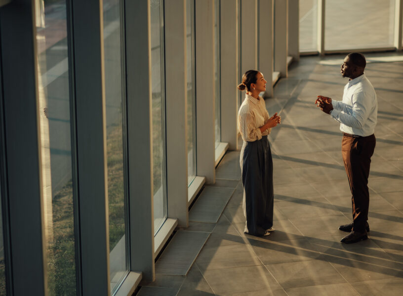 Businesspeople discussing near window in modern office building Two businesspeople are having a discussion near a large window in a modern office building, illuminated by warm sunlight