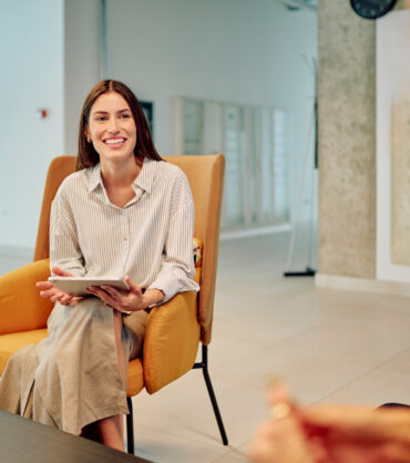 Businesswoman holding tablet and smiling while sitting on armchair and discussing with colleague in modern office